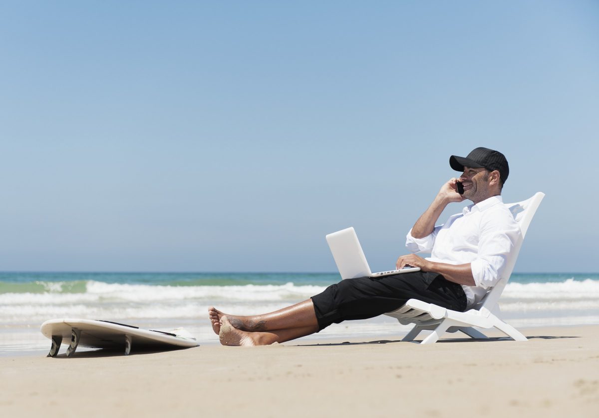 A Businessman Sits On A Beach Chair On The Beach Working On A Laptop Computer And Talking On The Phone With His Surfboard Sitting At His Feet; Tarifa Cadiz Andalusia Spain